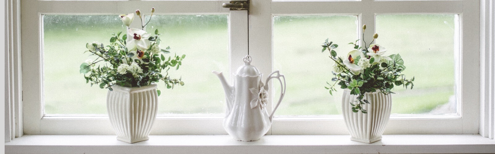 a white teapot surrounded by 2 pots of flowers on a window-sill.